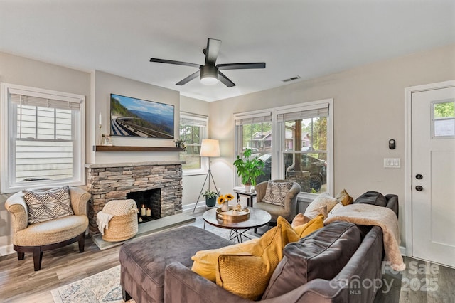 living area featuring a ceiling fan, a stone fireplace, visible vents, wood finished floors, and baseboards