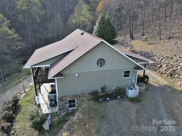 view of side of home with a forest view, central AC unit, stone siding, and roof with shingles