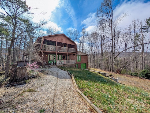 rear view of property featuring a gambrel roof, an attached garage, a wooden deck, gravel driveway, and a balcony