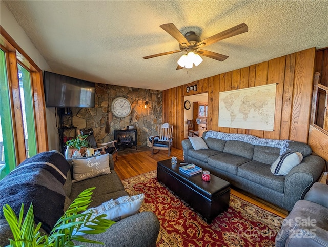 living area featuring wooden walls, wood finished floors, a wood stove, a textured ceiling, and ceiling fan