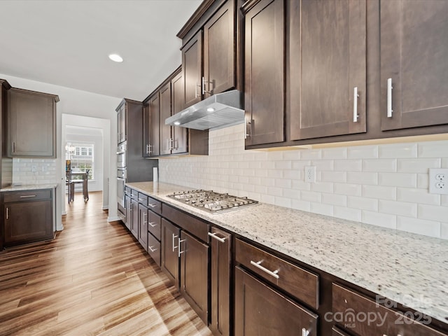 9582 Creighton Rd NW, Concord, NC 28027 - kitchen with light wood-type flooring, stainless steel appliances, under cabinet range hood, decorative backsplash, and dark brown cabinets