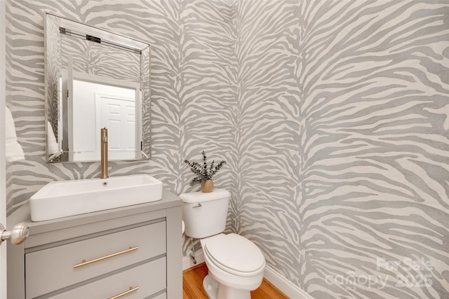 half bathroom with vanity, toilet, wallpapered walls, and wood finished floors