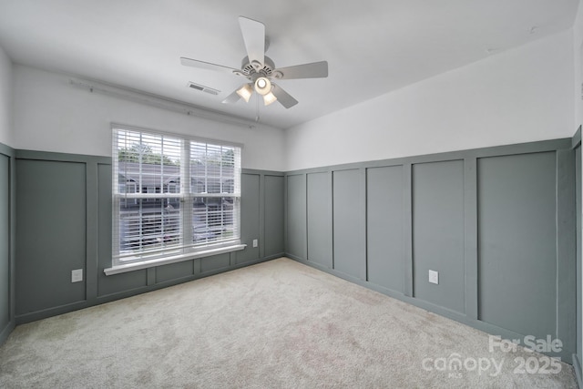 carpeted empty room with a decorative wall, visible vents, a ceiling fan, and wainscoting
