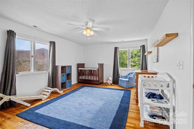 bedroom with a textured ceiling, visible vents, a crib, ceiling fan, and wood finished floors