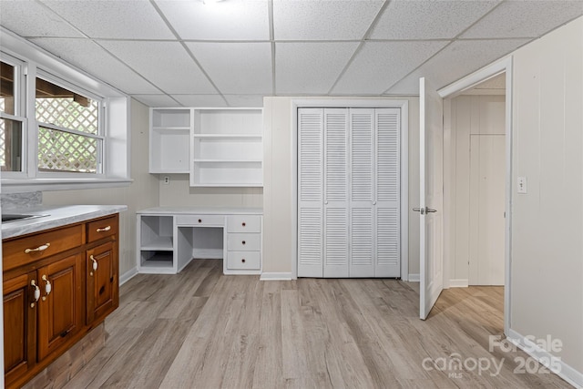 unfurnished office featuring baseboards, built in desk, a drop ceiling, and light wood finished floors