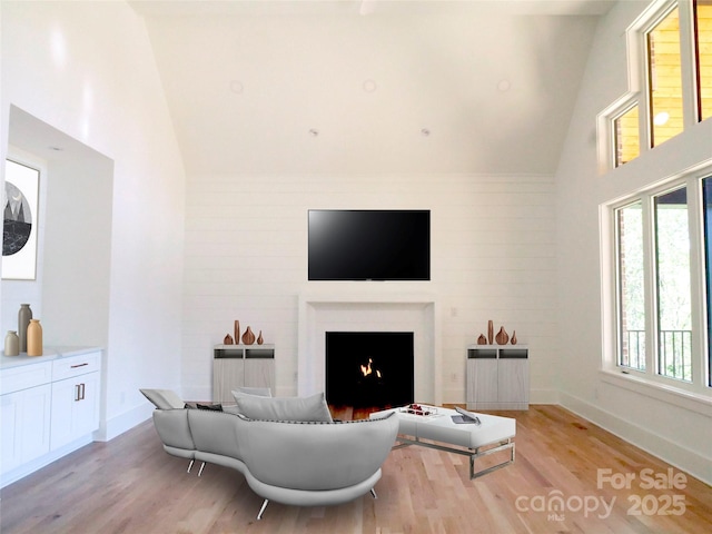 living room featuring light wood-style flooring, high vaulted ceiling, baseboards, and a lit fireplace