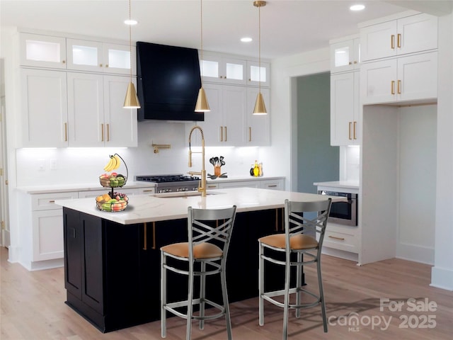 kitchen featuring ventilation hood, light wood finished floors, glass insert cabinets, and an island with sink