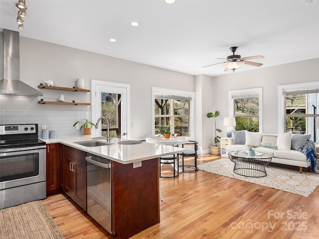 kitchen with open floor plan, light countertops, a sink, appliances with stainless steel finishes, and wall chimney exhaust hood
