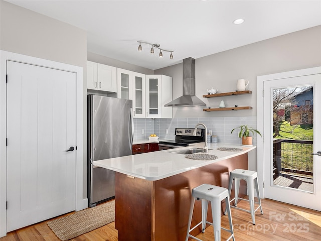 kitchen featuring a peninsula, wall chimney exhaust hood, appliances with stainless steel finishes, glass insert cabinets, and backsplash