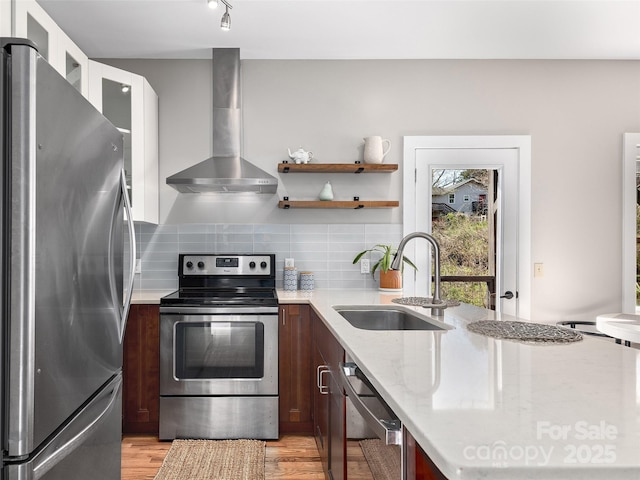 kitchen with decorative backsplash, light wood-style flooring, appliances with stainless steel finishes, a sink, and wall chimney range hood