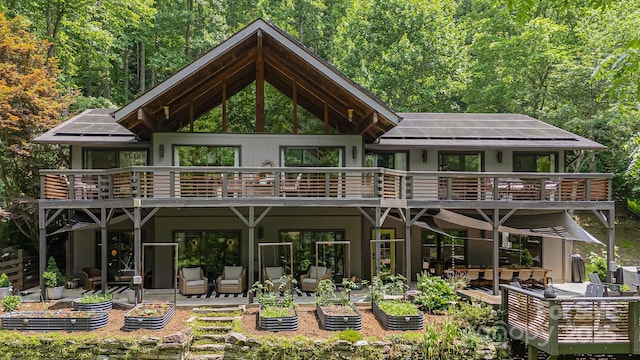 back of property featuring a wooded view, a balcony, outdoor dining space, a patio, and solar panels