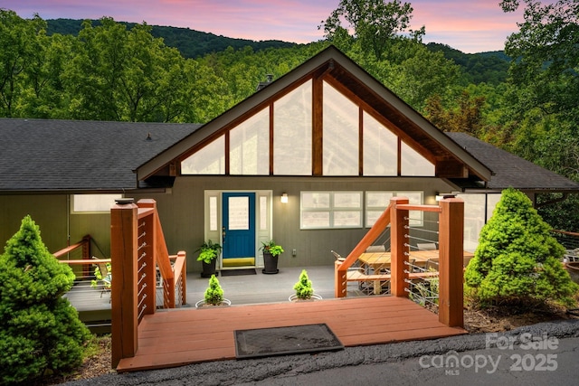 rear view of property with a wooden deck, roof with shingles, and a wooded view