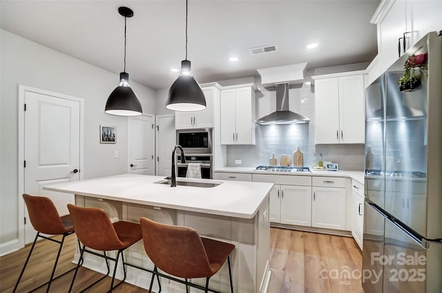kitchen featuring light wood finished floors, tasteful backsplash, appliances with stainless steel finishes, wall chimney range hood, and visible vents