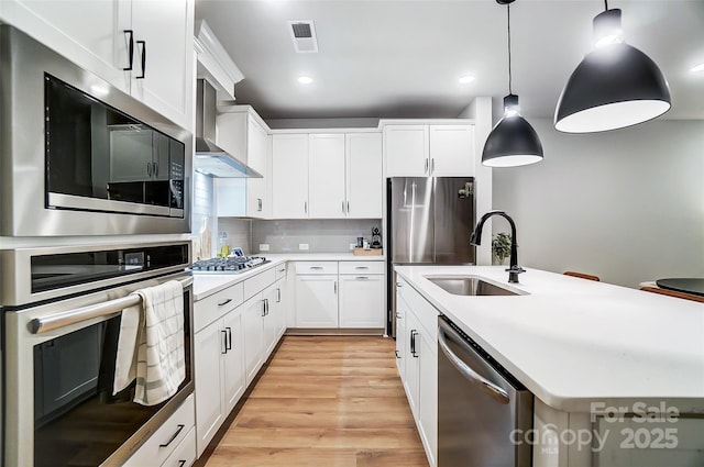 kitchen featuring a center island with sink, a sink, light wood-style floors, wall chimney range hood, and appliances with stainless steel finishes