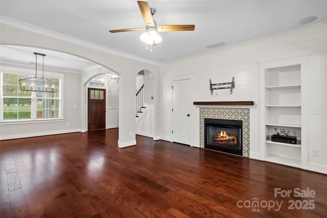unfurnished living room with ornamental molding, built in features, baseboards, ceiling fan, and a tile fireplace