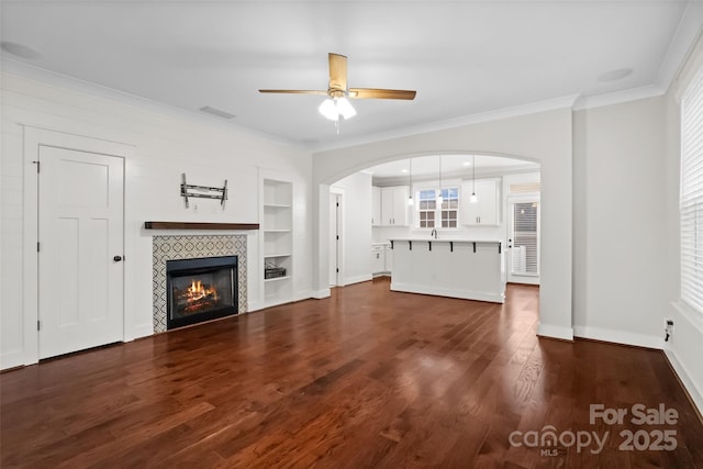unfurnished living room with ceiling fan, built in features, arched walkways, dark wood finished floors, and ornamental molding