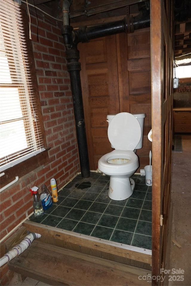 bathroom with toilet, plenty of natural light, tile patterned floors, and brick wall