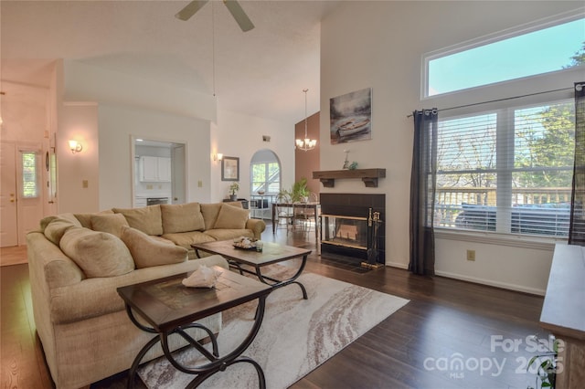 living area featuring ceiling fan with notable chandelier, high vaulted ceiling, dark wood-type flooring, baseboards, and a tile fireplace