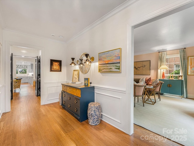 hall featuring light wood finished floors, wainscoting, crown molding, and a decorative wall
