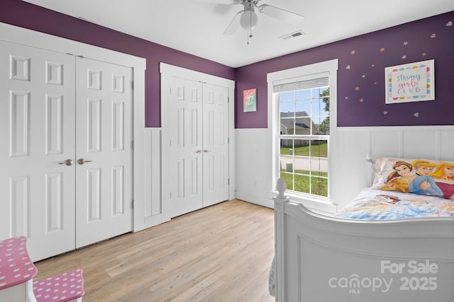bedroom with light wood finished floors, a ceiling fan, multiple closets, visible vents, and wainscoting