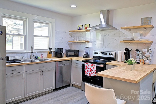 kitchen featuring open shelves, wooden counters, a sink, appliances with stainless steel finishes, and wall chimney exhaust hood