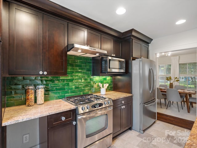 kitchen featuring light stone countertops, dark brown cabinets, under cabinet range hood, tasteful backsplash, and stainless steel appliances
