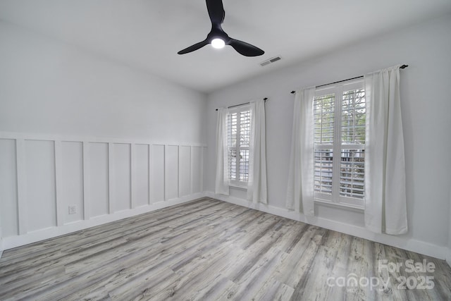 unfurnished room featuring visible vents, a decorative wall, ceiling fan, wainscoting, and wood finished floors