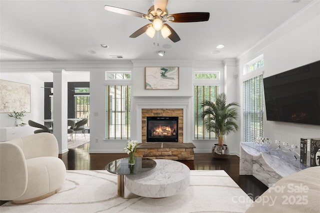 living room featuring crown molding, a ceiling fan, visible vents, a stone fireplace, and wood finished floors