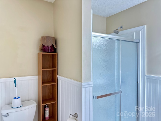full bath featuring toilet, a stall shower, a textured ceiling, and a wainscoted wall