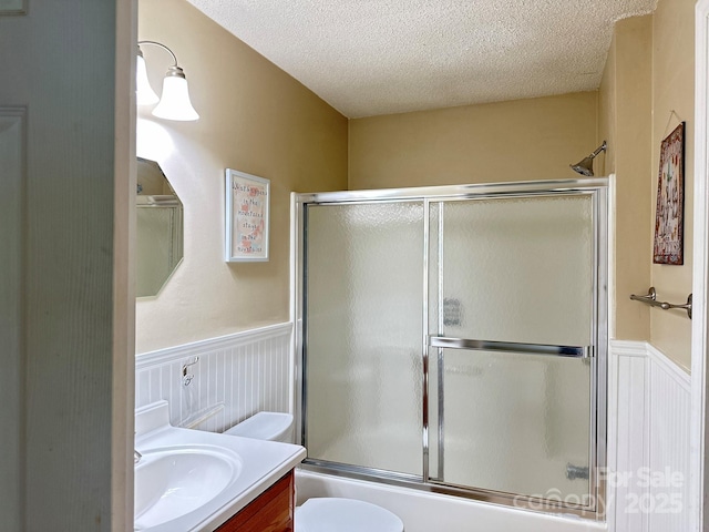 bathroom with a textured ceiling, vanity, a wainscoted wall, toilet, and bath / shower combo with glass door