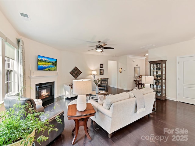 living room featuring a glass covered fireplace, ceiling fan, baseboards, dark wood-style flooring, and visible vents