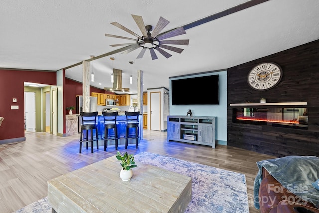 living room with baseboards, ceiling fan, light wood finished floors, a glass covered fireplace, and lofted ceiling