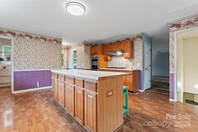 kitchen with light wood-style floors, gas cooktop, a center island, under cabinet range hood, and wallpapered walls