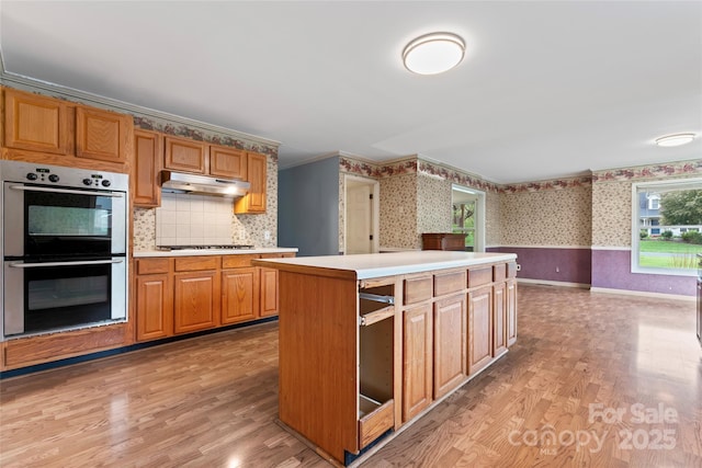 kitchen featuring light countertops, light wood-type flooring, under cabinet range hood, wallpapered walls, and stainless steel double oven