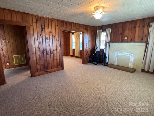 living area featuring visible vents, wooden walls, a brick fireplace, carpet, and a ceiling fan