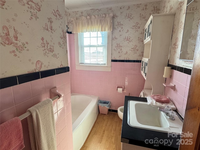 bathroom featuring tile walls, wainscoting, a garden tub, wallpapered walls, and wood finished floors