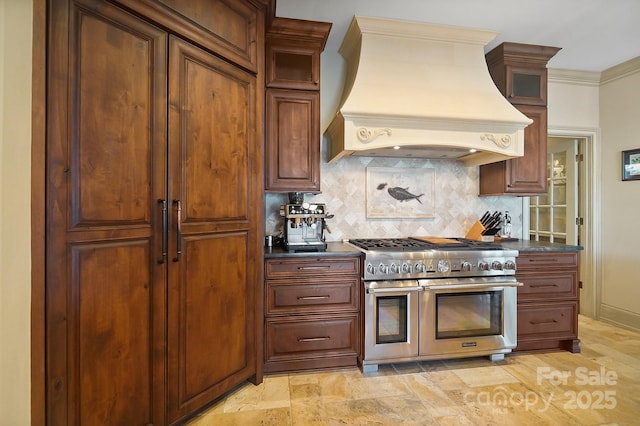 kitchen featuring custom exhaust hood, glass insert cabinets, dark countertops, backsplash, and range with two ovens