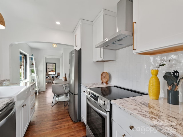 kitchen featuring arched walkways, wall chimney range hood, white cabinetry, and stainless steel appliances