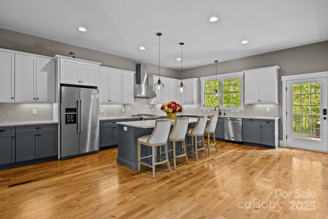 kitchen featuring wall chimney range hood, appliances with stainless steel finishes, tasteful backsplash, light wood-style floors, and a breakfast bar