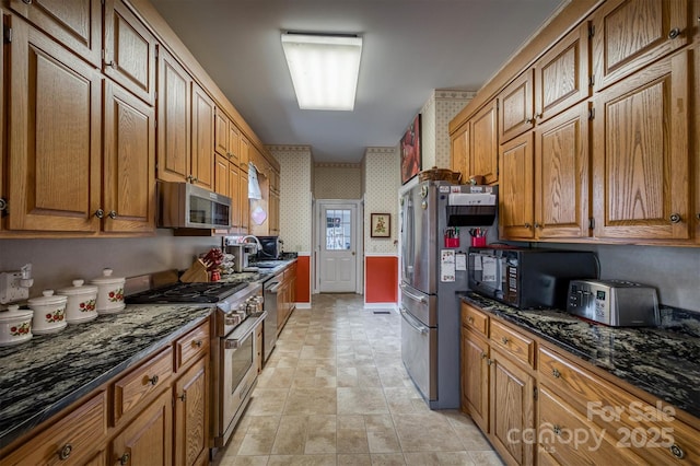 kitchen featuring wallpapered walls, a toaster, brown cabinetry, dark stone counters, and stainless steel appliances