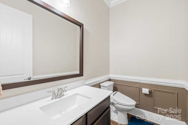 bathroom with a decorative wall, vanity, ornamental molding, a wainscoted wall, and toilet
