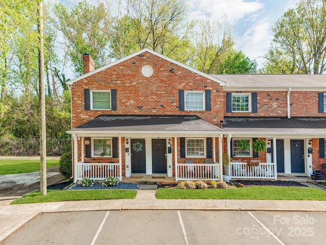 view of front of property with a shingled roof, a porch, uncovered parking, a chimney, and brick siding