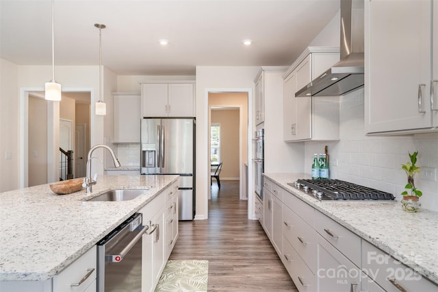 kitchen featuring light wood-style floors, wall chimney range hood, appliances with stainless steel finishes, a sink, and a center island with sink