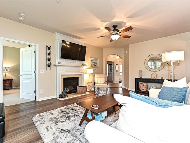 living room featuring a glass covered fireplace, a ceiling fan, baseboards, arched walkways, and wood finished floors