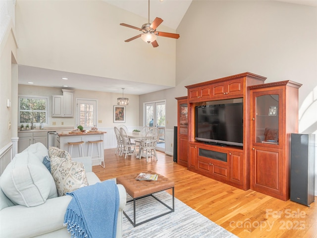 living room featuring light wood finished floors, ceiling fan with notable chandelier, a wainscoted wall, and a healthy amount of sunlight