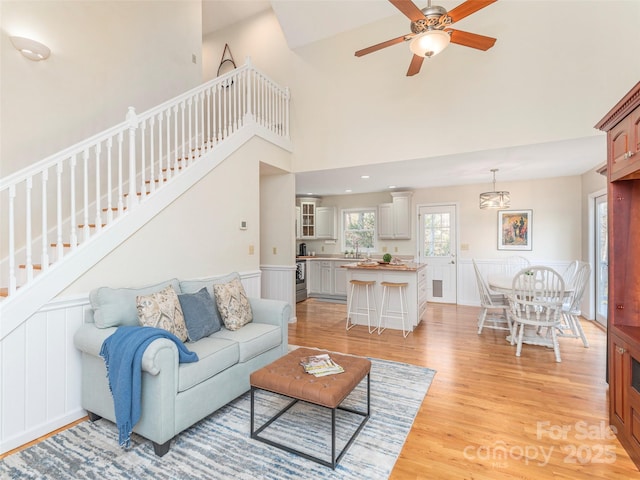 living area featuring stairway, light wood-style flooring, a high ceiling, a ceiling fan, and a wainscoted wall