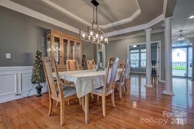 dining room with crown molding, a tray ceiling, ornate columns, light wood finished floors, and a wainscoted wall