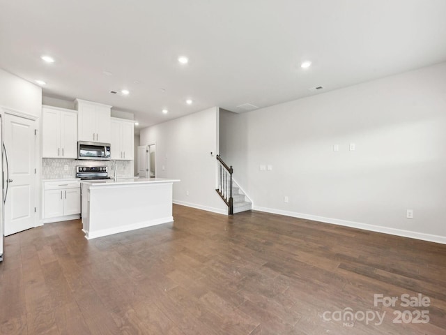 kitchen featuring appliances with stainless steel finishes, decorative backsplash, dark wood finished floors, a center island with sink, and baseboards