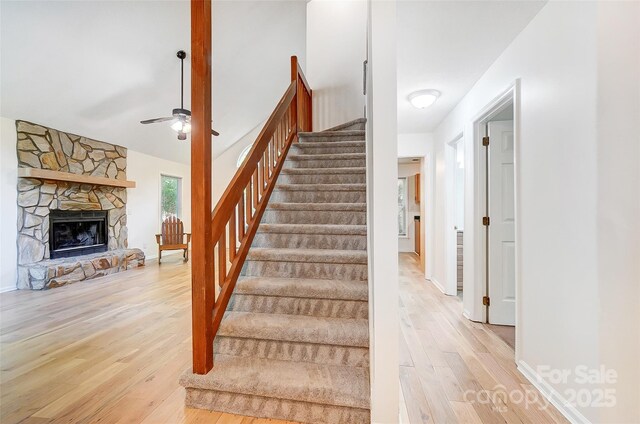stairway with wood finished floors, baseboards, a ceiling fan, and a stone fireplace