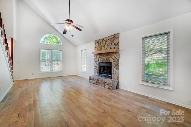unfurnished living room featuring a stone fireplace, ceiling fan, light wood finished floors, stairway, and high vaulted ceiling